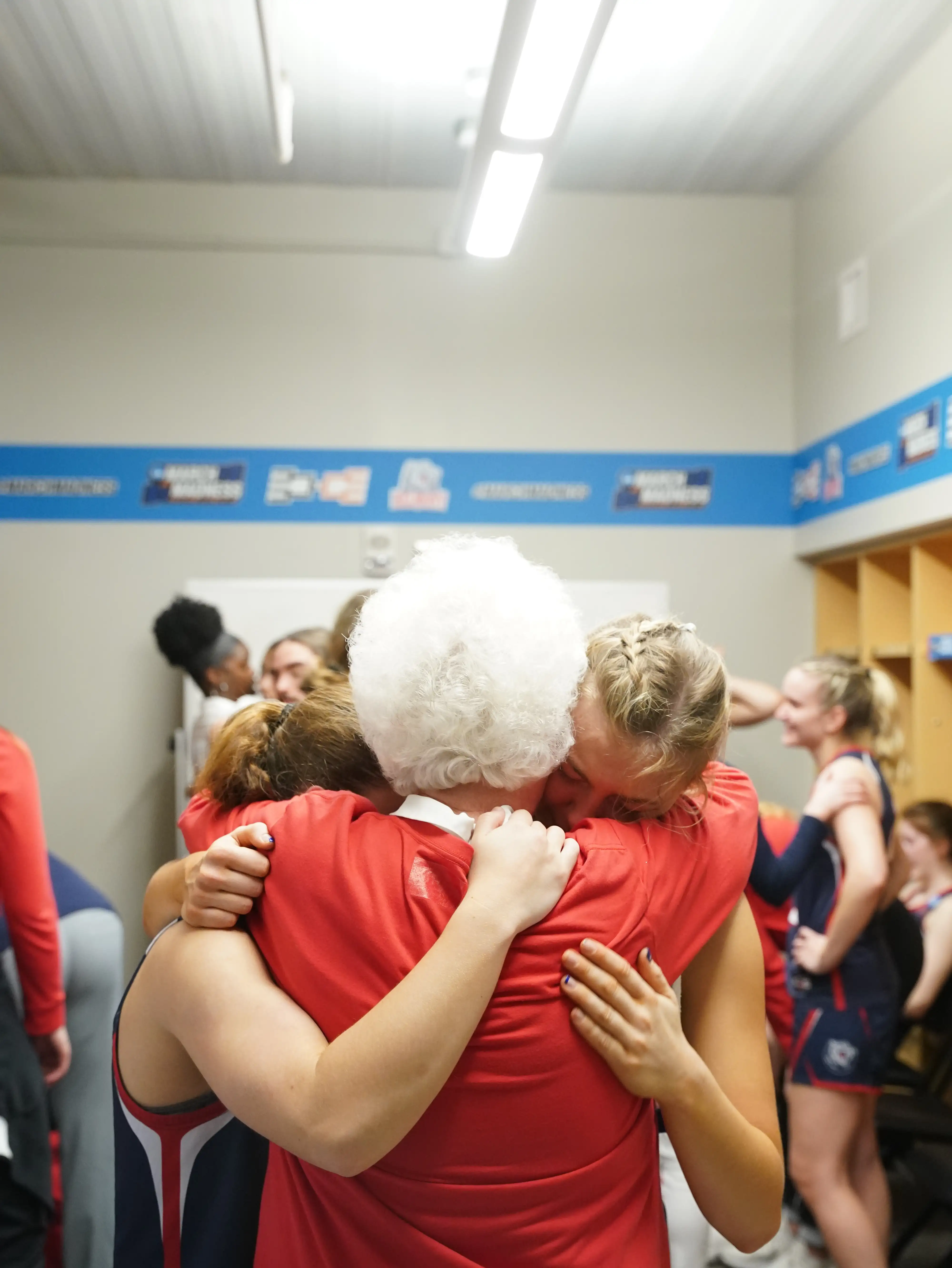 Belmont Women’s Basketball players embrace longtime coach and mentor Betty Wiseman in an emotional group hug in the locker room. Wiseman, with white hair and a red shirt, is surrounded by players in uniform.