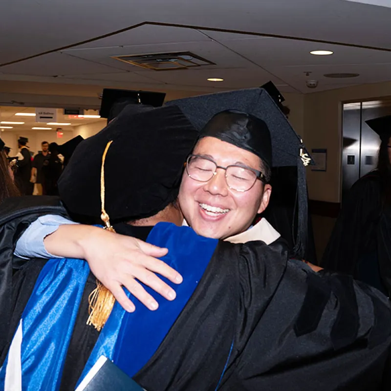 A graduate in cap and gown hugging a faculty member during a graduation ceremony.