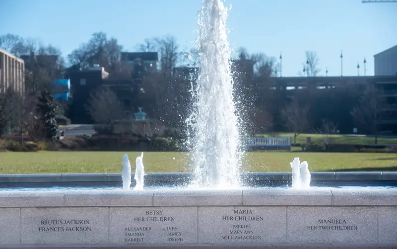 Freedom Plaza fountain showing wall engraved with family names beneath a large central water fountain jet and two smaller side jets, framed by a green lawn and campus buildings under a clear blue sky.
