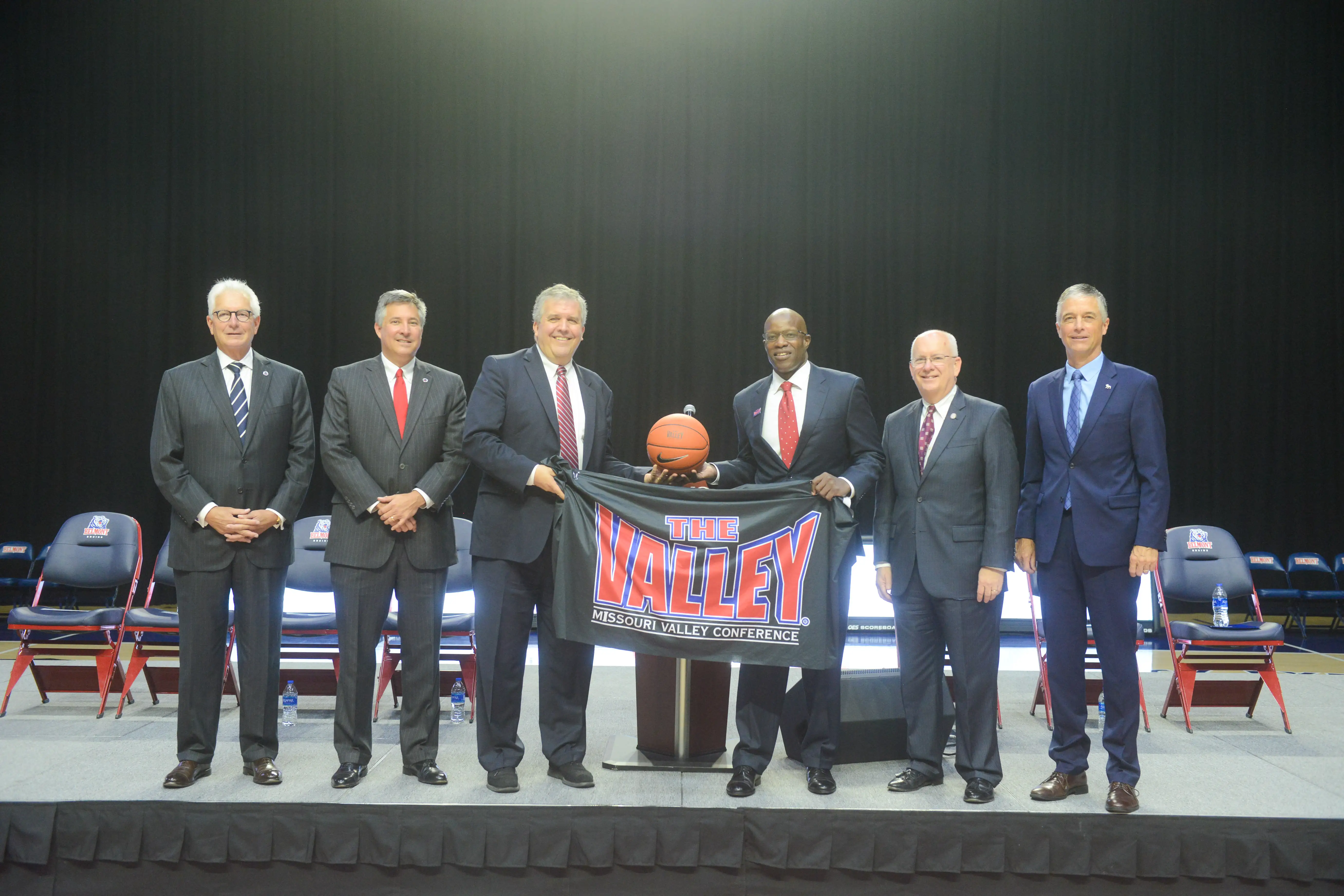 Six men in suits, including President Dr. Greg Jones, stand on a stage in front of empty chairs, posing for a photo. Two of them hold a basketball and a black banner that reads “THE VALLEY: MISSOURI VALLEY CONFERENCE,” signifying Belmont University’s move to the MVC.