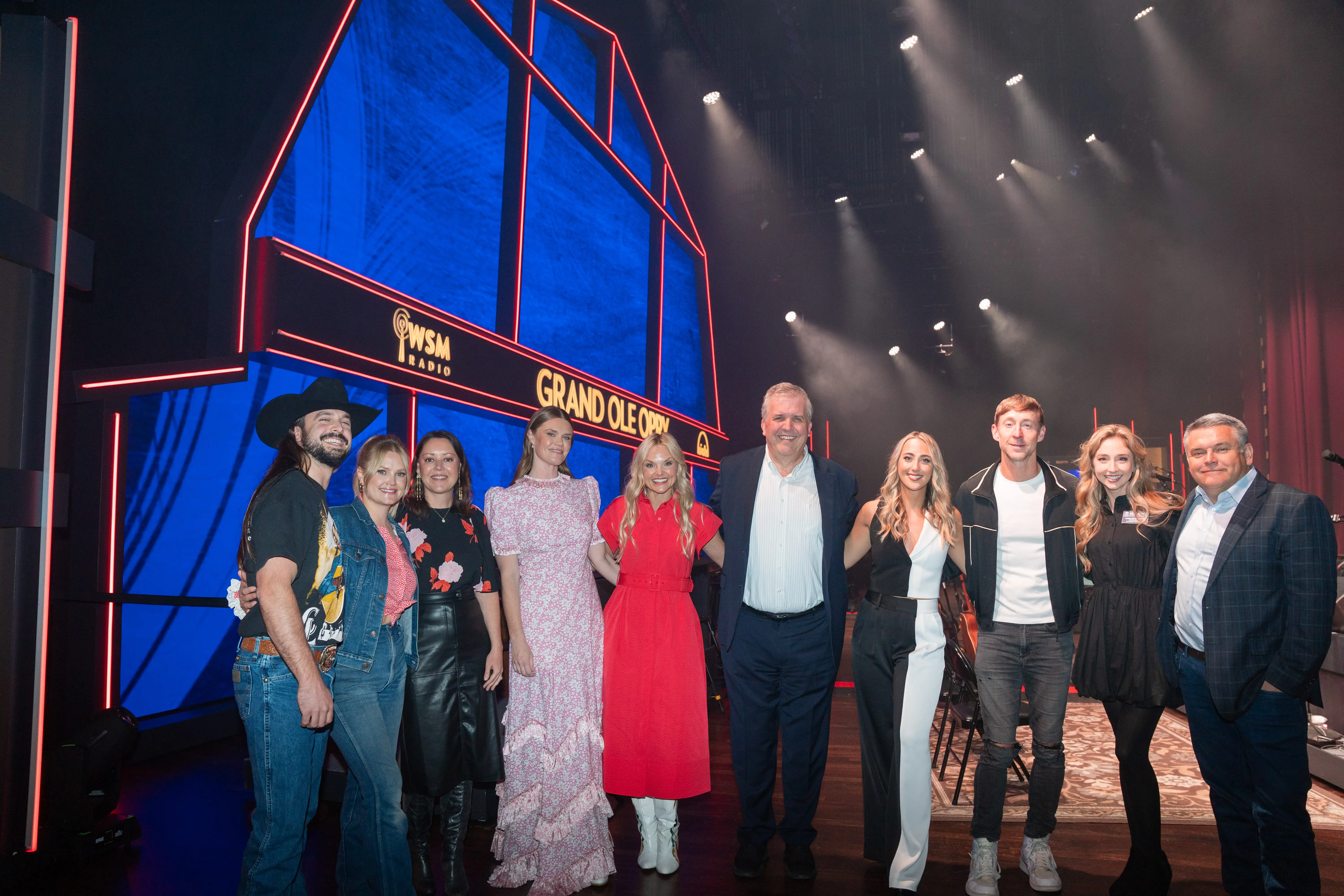 A group of Belmont University-affiliated individuals, including staff and alumni, pose on stage at the Grand Ole Opry in front of a brightly lit red and blue Opry backdrop.