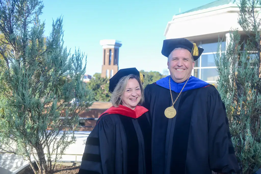 An outdoor photo of Dr. Greg Jones and Rev. Susan Pendleton Jones smiling, both in caps and gowns on the day of Dr Jones' Inauguration with the Bell Tower in the background.