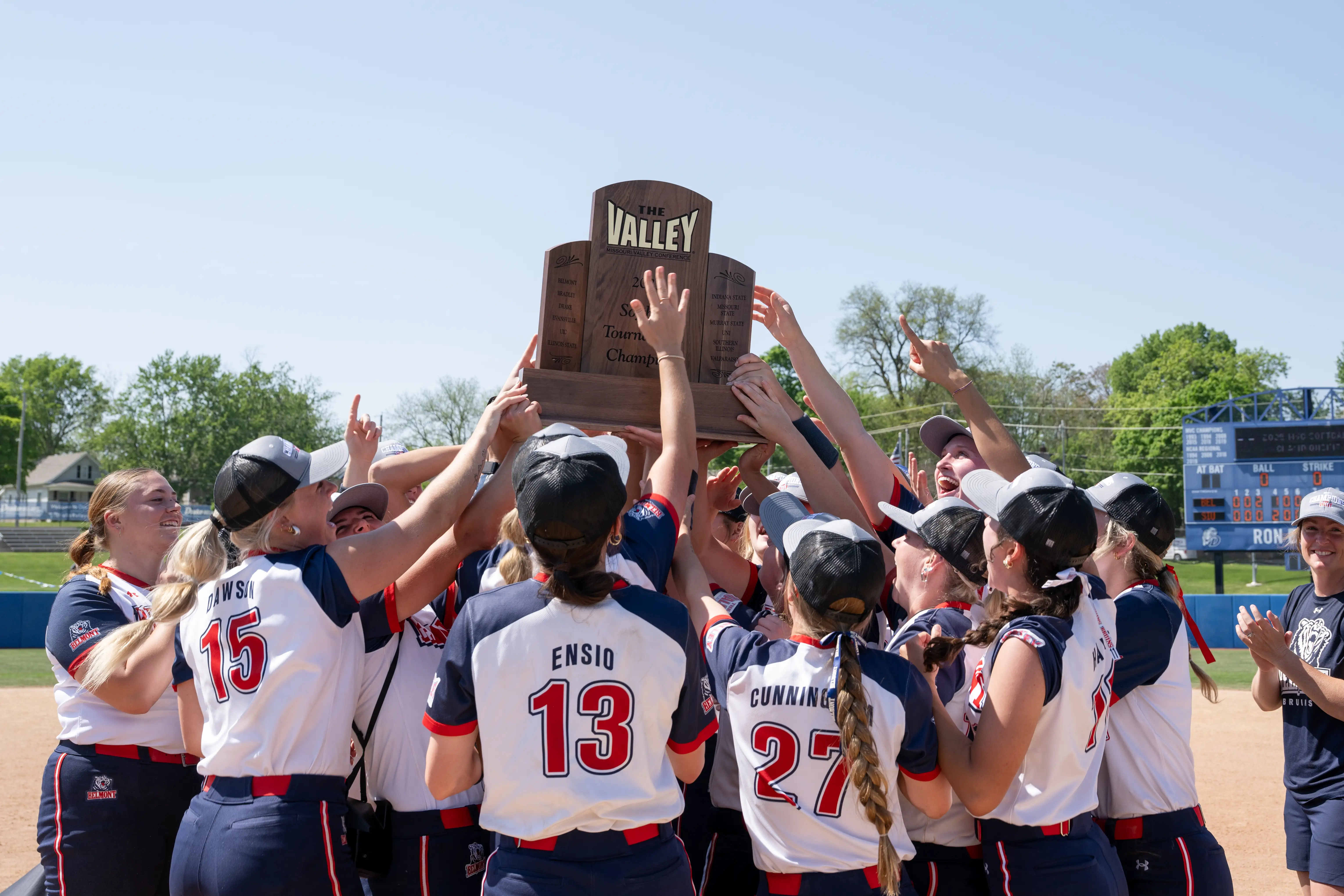 The Belmont University softball team huddles together on the field, joyfully raising a championship trophy.