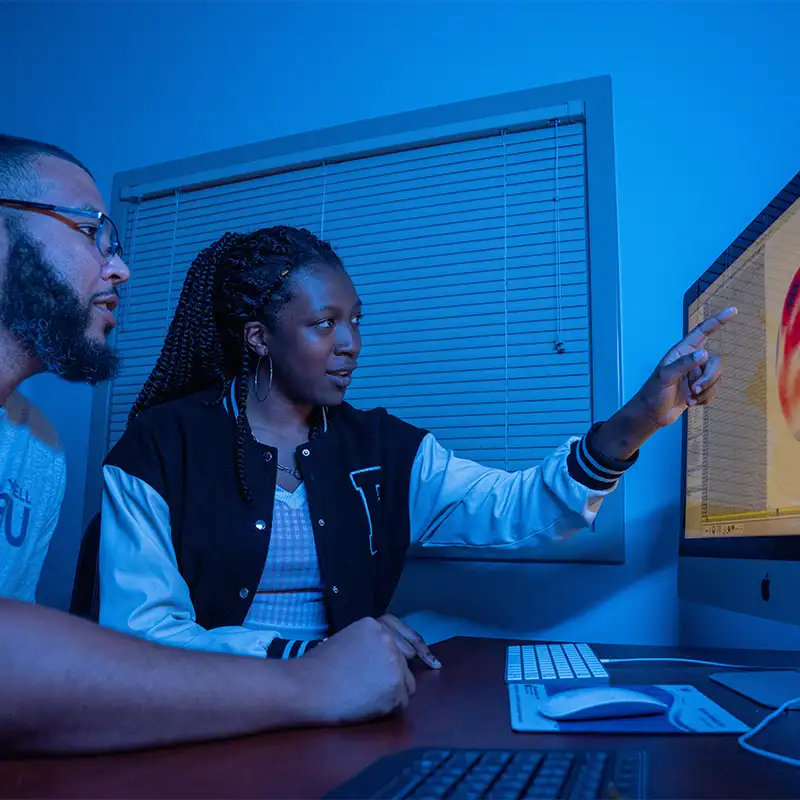 Two people seated at a desk in a darkened room bathed in blue light: a young Black woman wearing a varsity-style jacket points at a colorful 3D graphic on a large desktop monitor while a bearded Black man in glasses watches attentively.