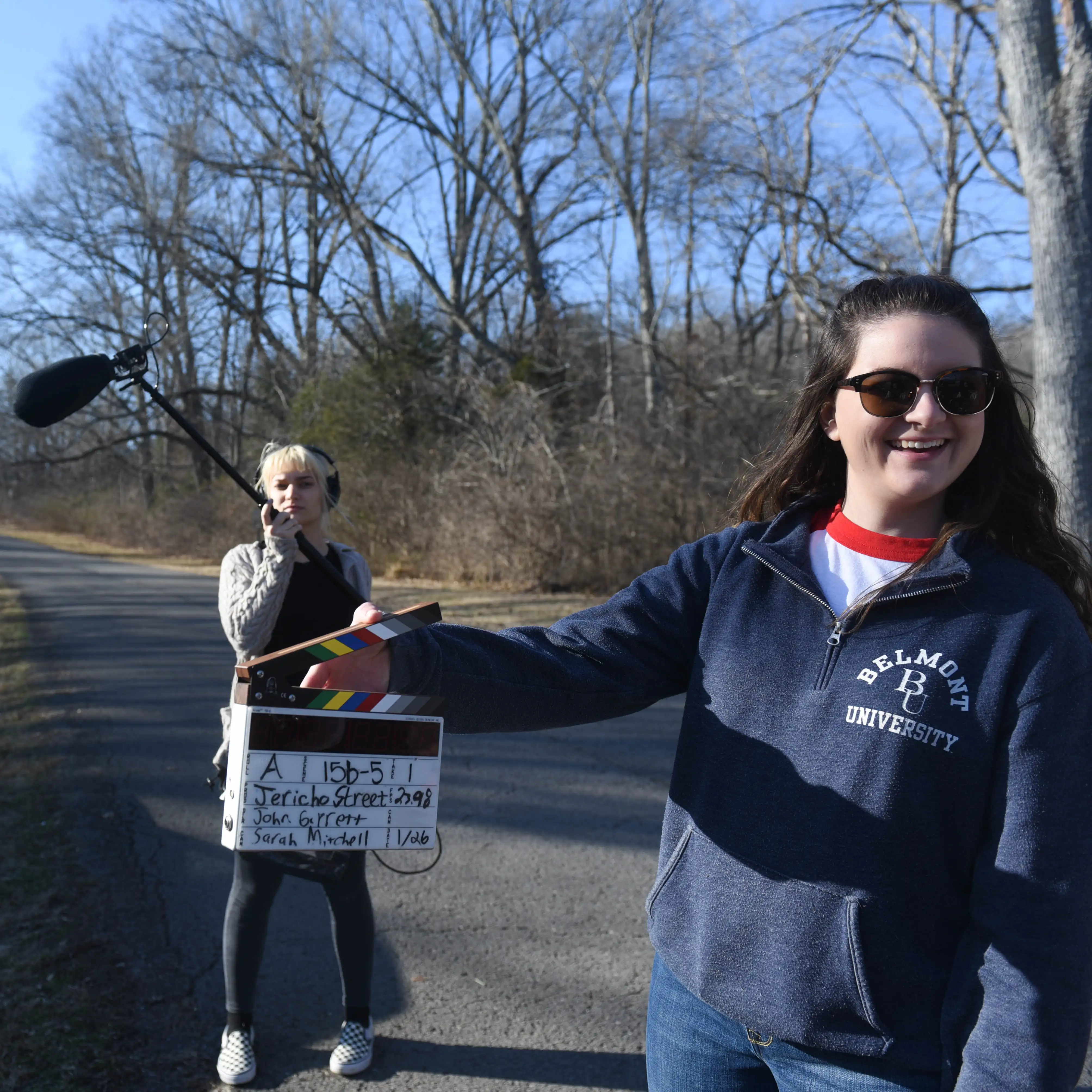 Belmont student smiles while holding a clapperboard on a rural road as another student operates a boom mic during a film production scene.