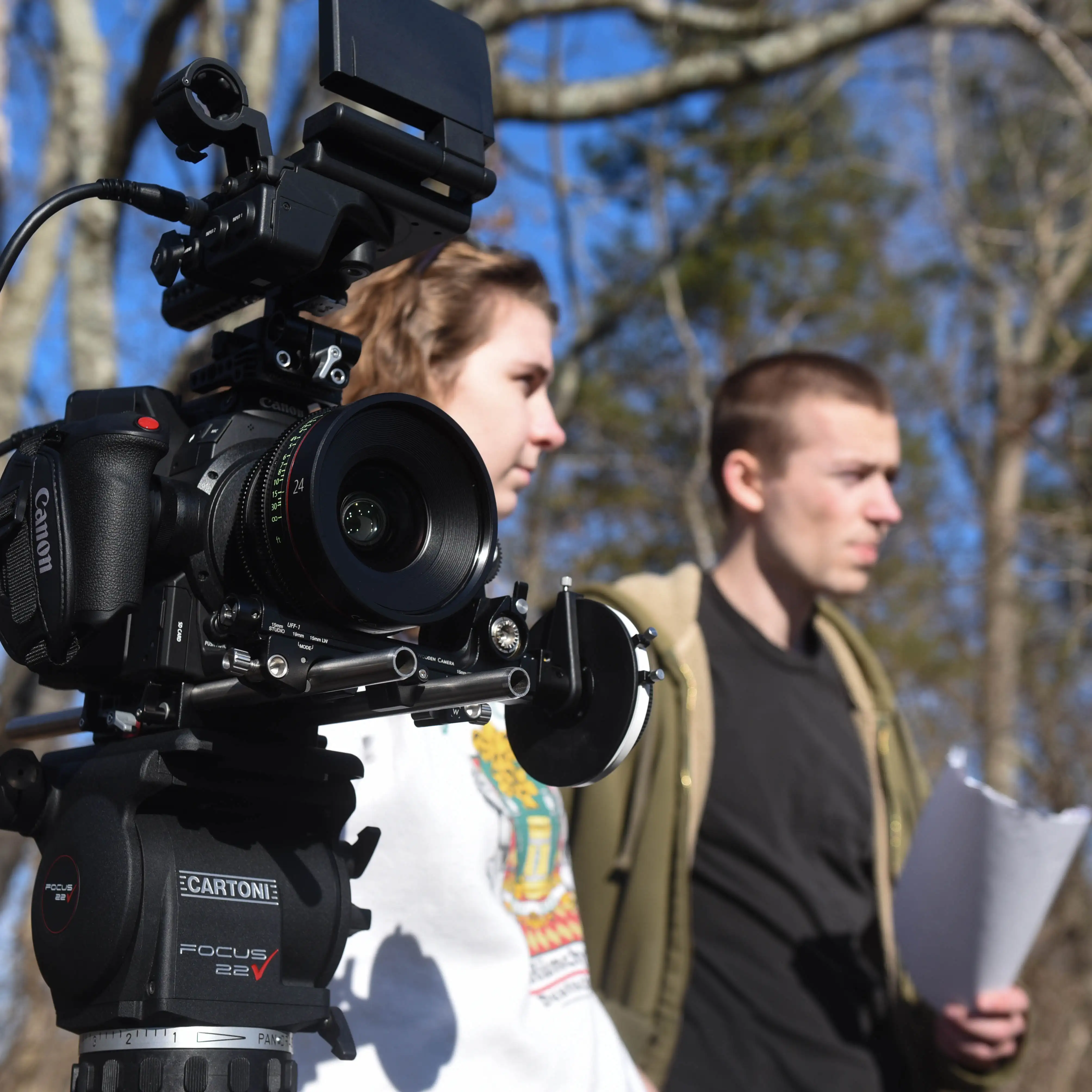 Close-up of a professional Canon camera on a tripod outdoors with two students blurred in the background, one holding a script during a film shoot.