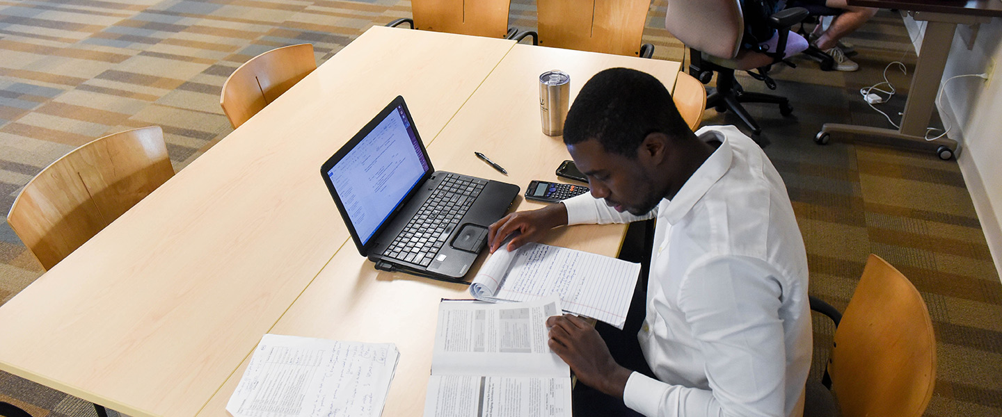 A male student sits at a table with a text book, laptop, and notes spread out in front of him.