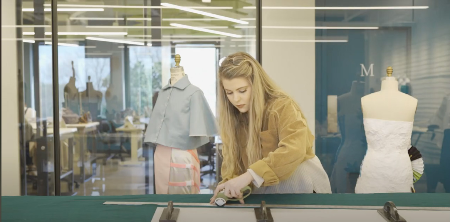 A screenshot from a video about Omore College at Belmont University. A female student stands at a table and works on a garment.