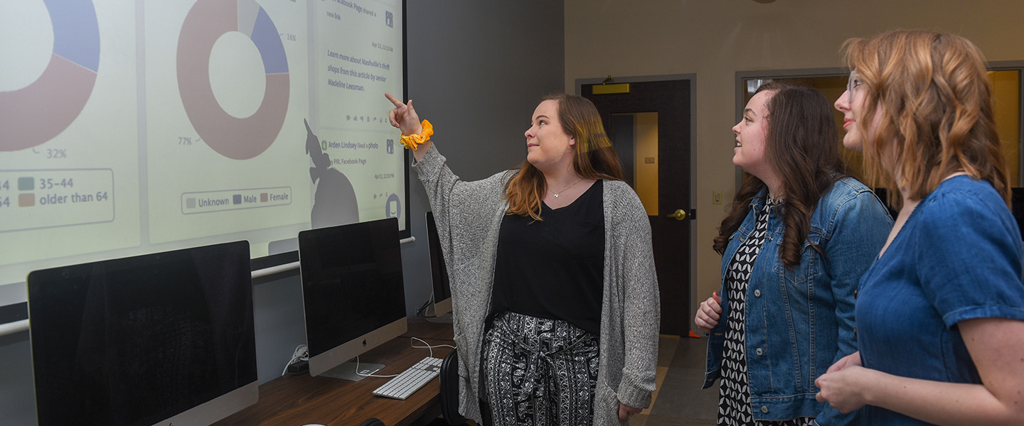 Three female students stand in front of a projection and point to a graph.