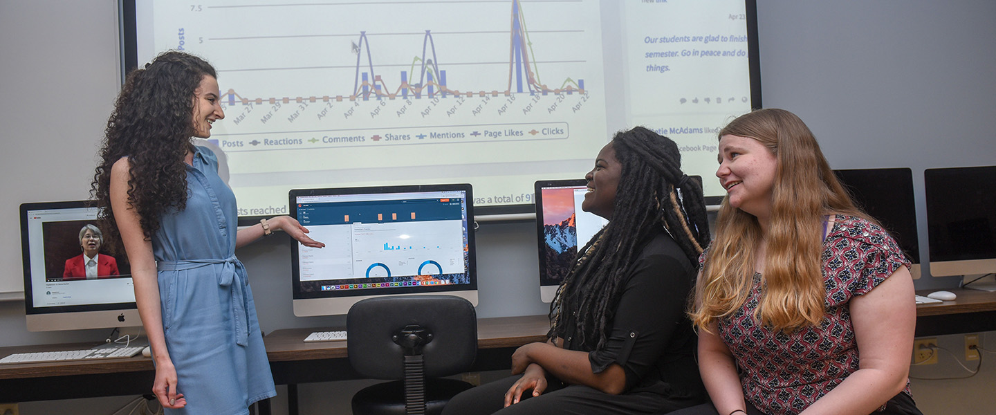 Three female students sit in a computer lab and point to charts displayed on computer monitors.