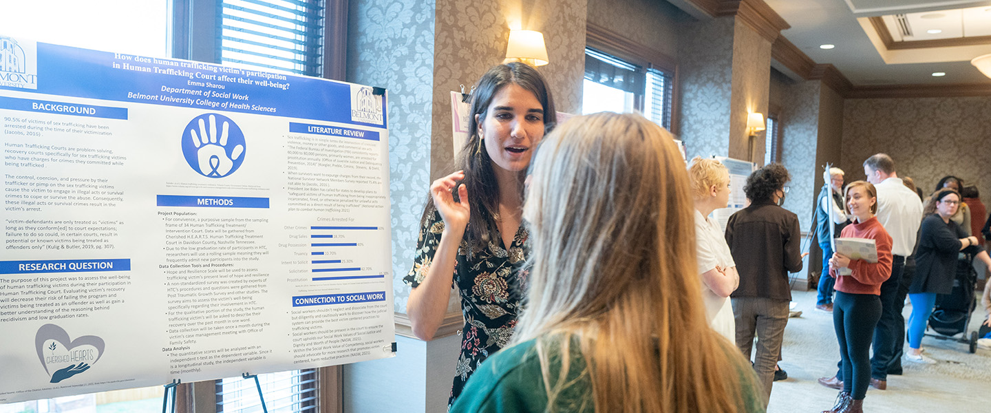 A female student presents her research on a poster board in a conference room.