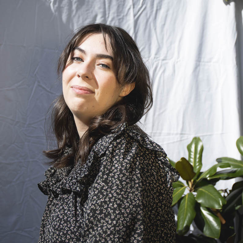 Art History alumna Gwyneth Cunningham sits in front of a white sheet next to a plant