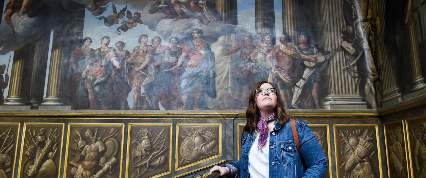 Student stands in front of historic art on wall on a study abroad experience