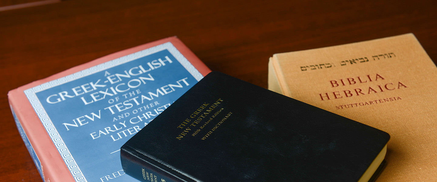 Three books on greek language, the new testament and the hebrew bible are spread across a table.