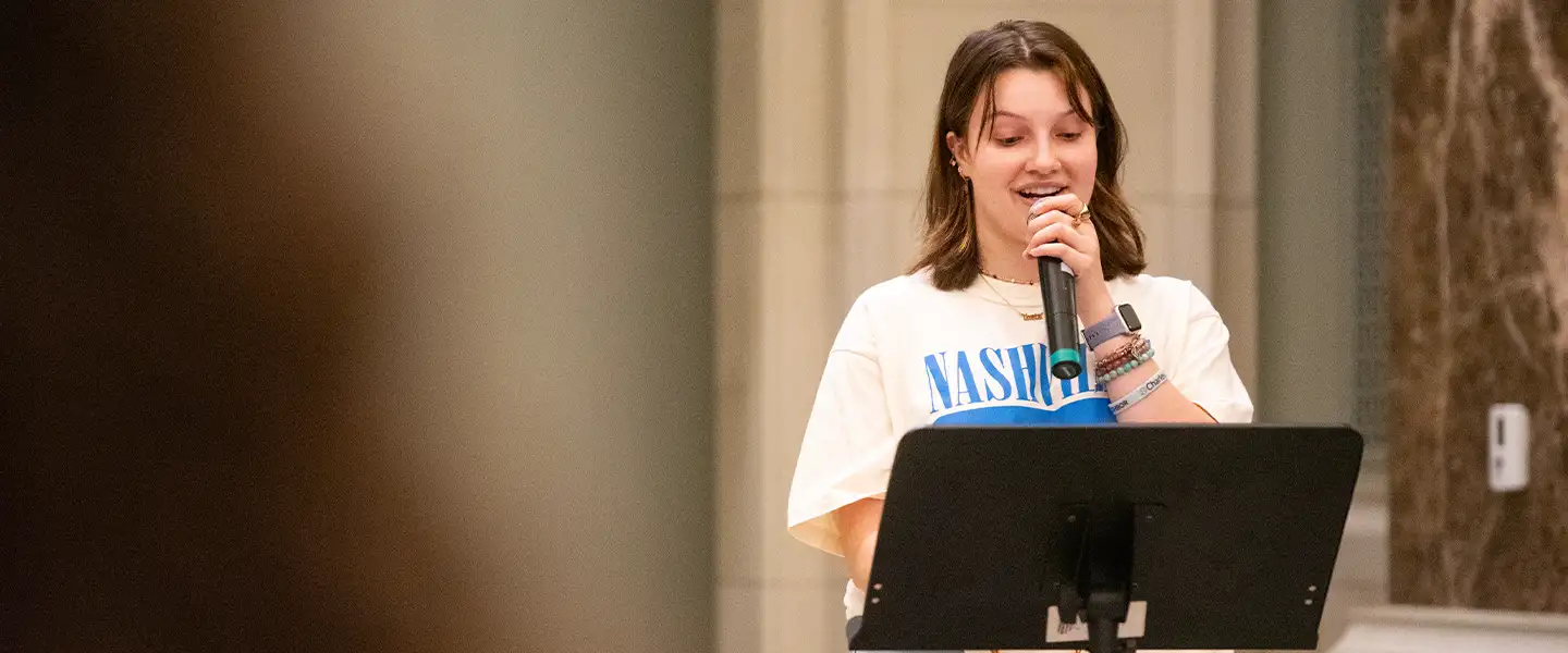 A young woman speaking into a microphone while standing behind a music stand, wearing a white shirt with 'NASHVILLE' printed in blue.