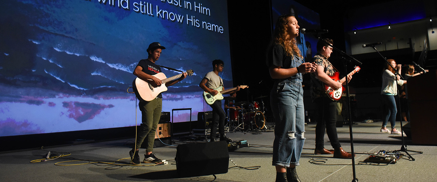 A female student stands in front of a microphone and leads worship with a band.