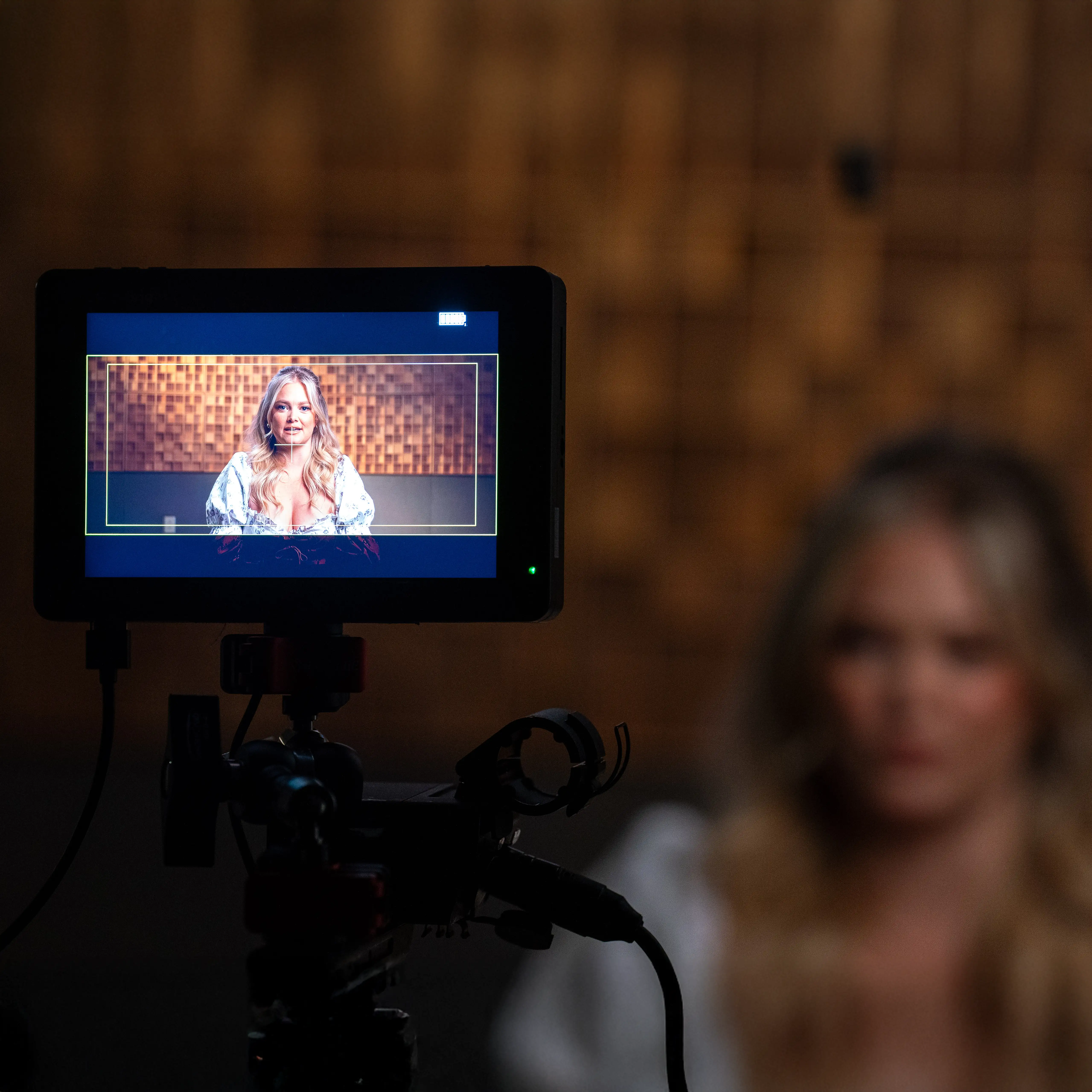 Camera monitor displays a woman on screen in a recording studio while she sits blurred in the background during a video shoot.