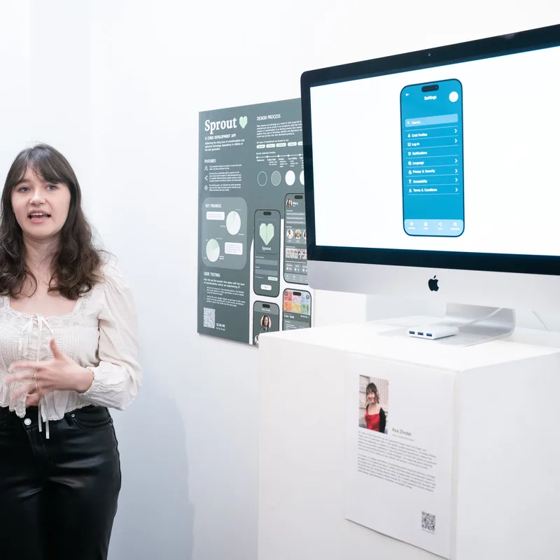A Watkins College of Art student stands in a white gallery space, explaining her mobile app design project ‘Sprout’ displayed on a printed poster board and an iMac showing the app’s interface.