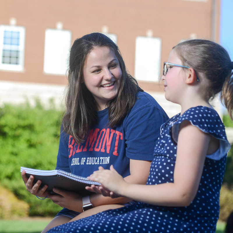 A woman in a Belmont T-Shirt sits with a young girl and reads to her, smiling.