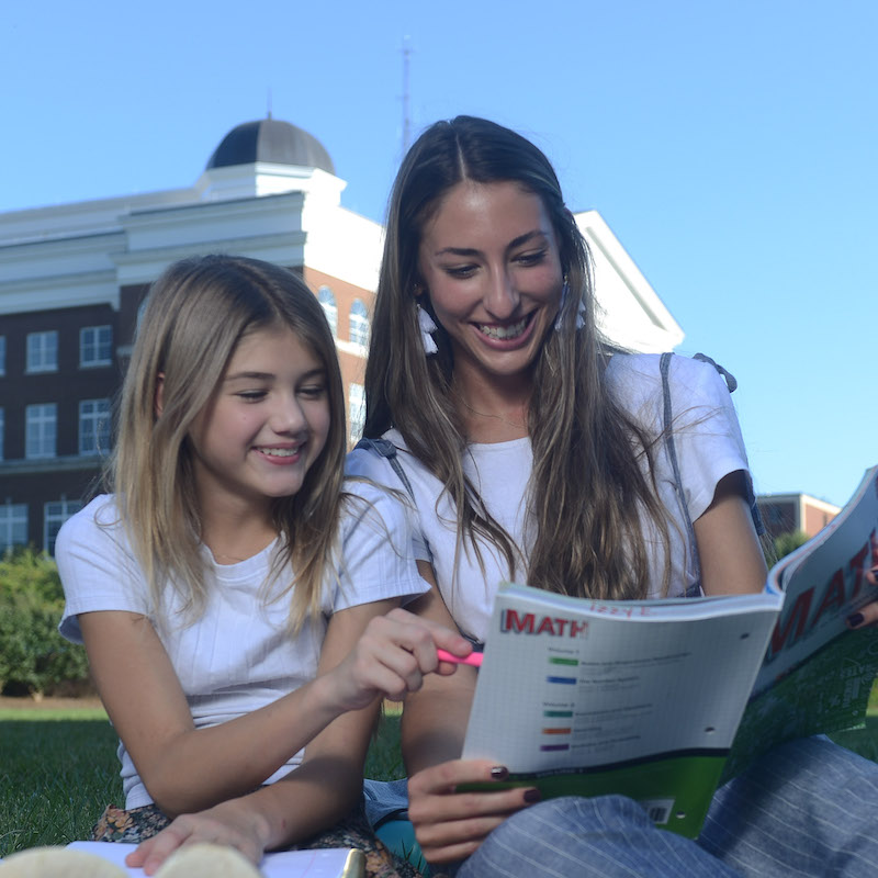 A female student sits on the lawn and reads to an elementary age child.
