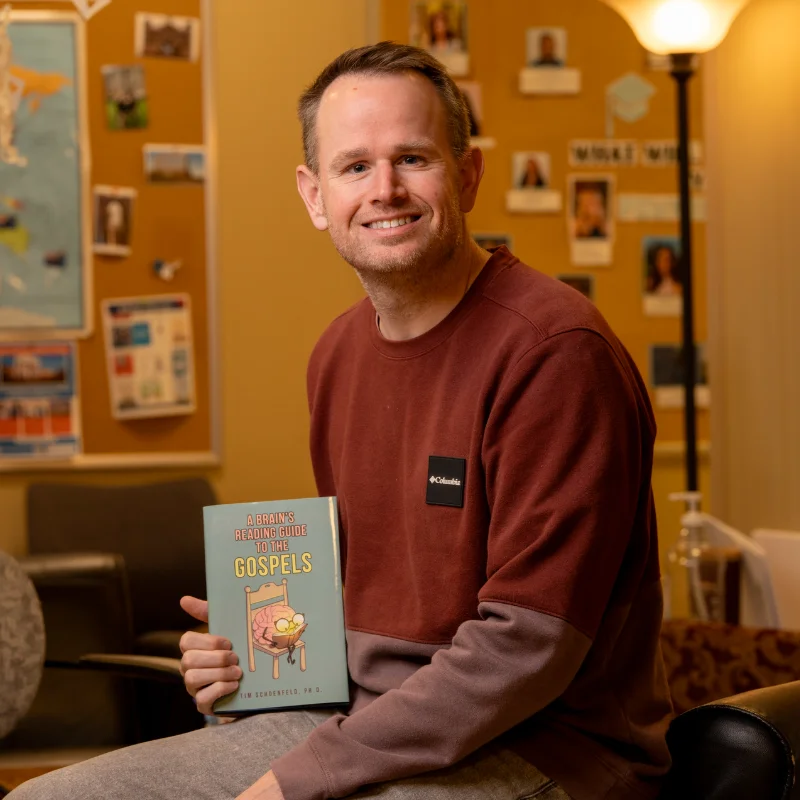 Psychology & Neurology Professor Tim Schoenfeld holding his book titled 'A Brain's Reading Guide to the Gospels', ideal for Christian studies.