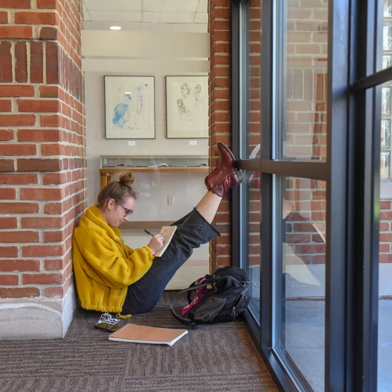 Focused student in yellow jacket writing in notebook on floor by a large window with artwork.