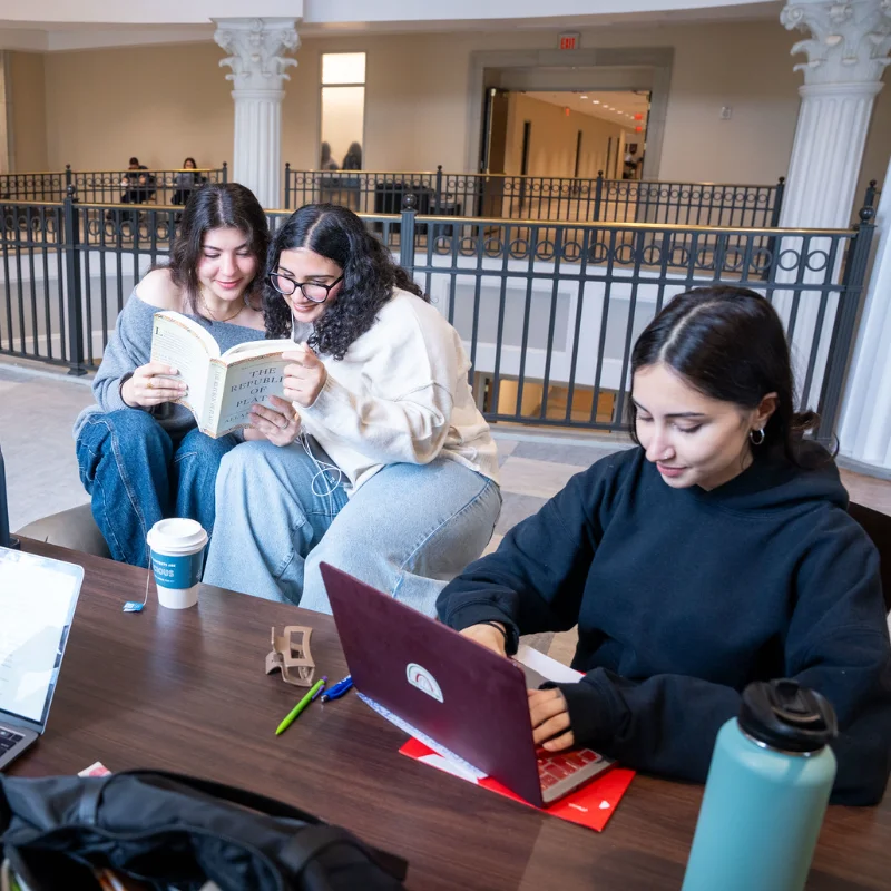 Two college students read a book while another works on a laptop.