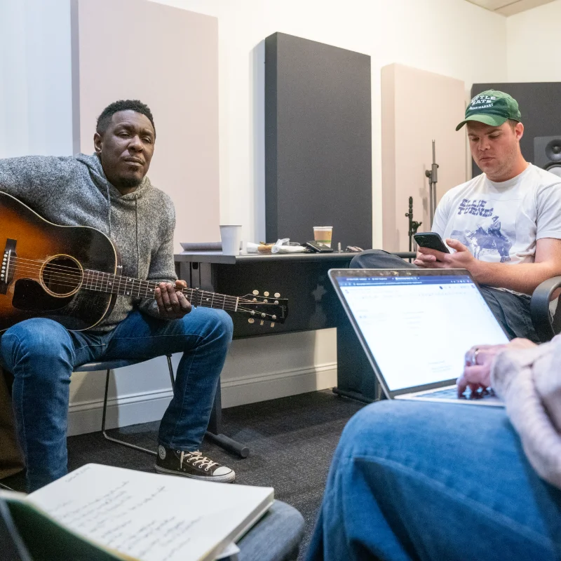Musician playing acoustic guitar during a studio session, with collaborators on laptops and phones in a recording room.