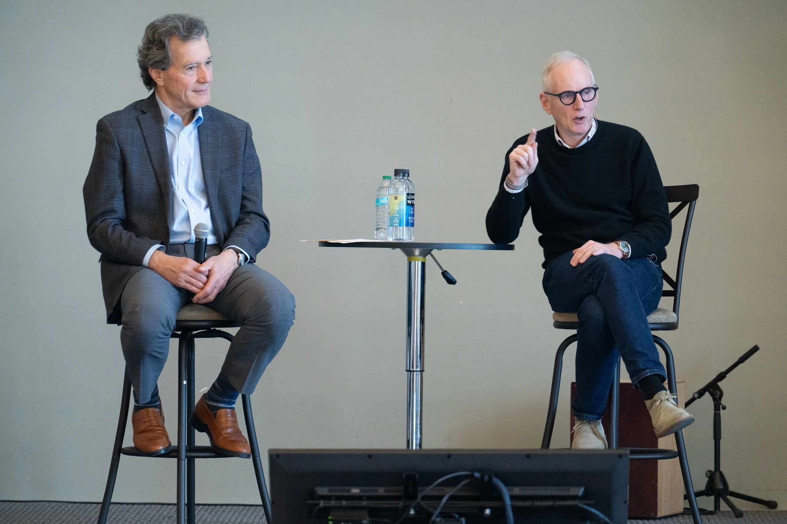Two men seated on stools on a stage during a discussion, one gesturing while speaking, the other listening.