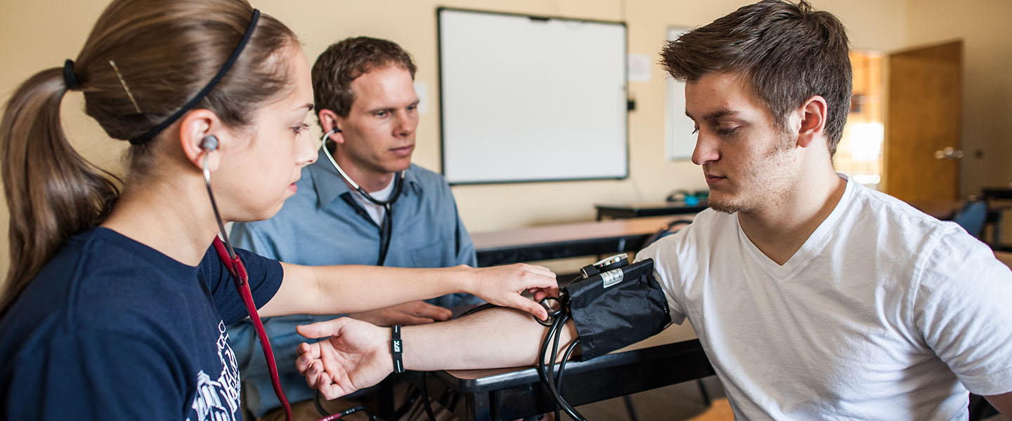 An exercise science student takes the blood pressure of another student with a professor in lab