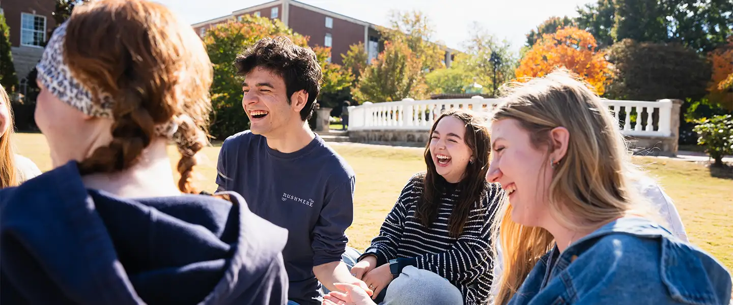 A group of Belmont students laughing and enjoying each other's company outdoors on the main lawn, with trees and a campus buildings in the background.