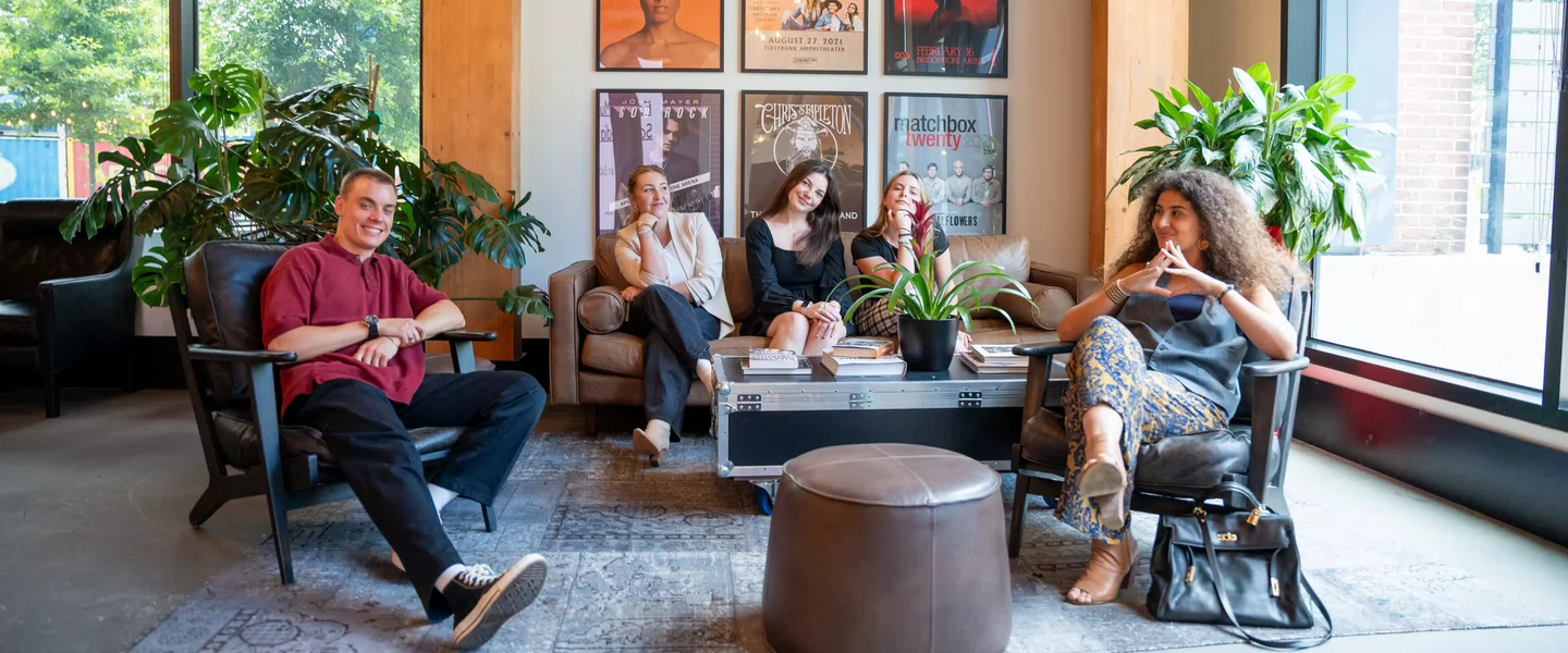 Five people sit and smile in a modern lounge area decorated with music posters, plants, and natural light from large windows, creating a relaxed and creative atmosphere.