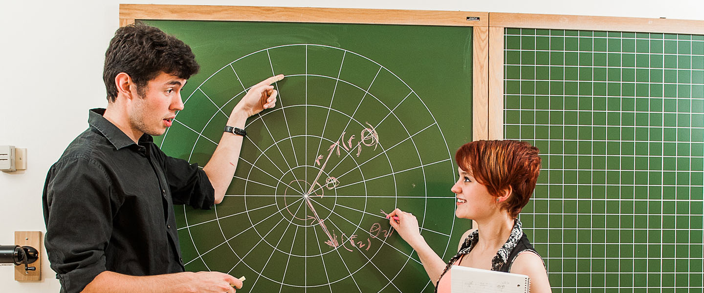 Two students stand in front of a green chalkboard with a math formula on it.