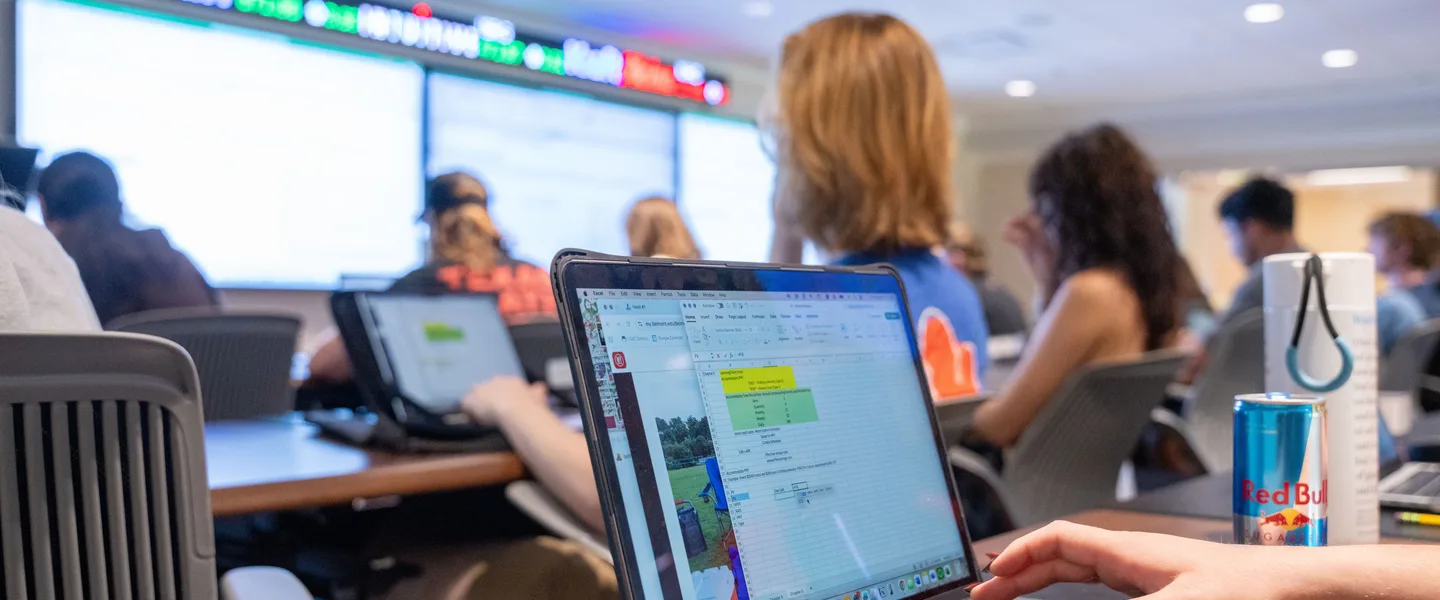 A classroom full of students working on laptops, with a large digital display board at the front showing data. The foreground shows a close-up of a student’s laptop screen displaying a spreadsheet.. 