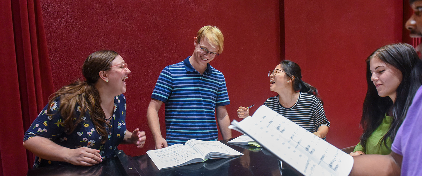 A group of students stand around a grand piano and laugh.