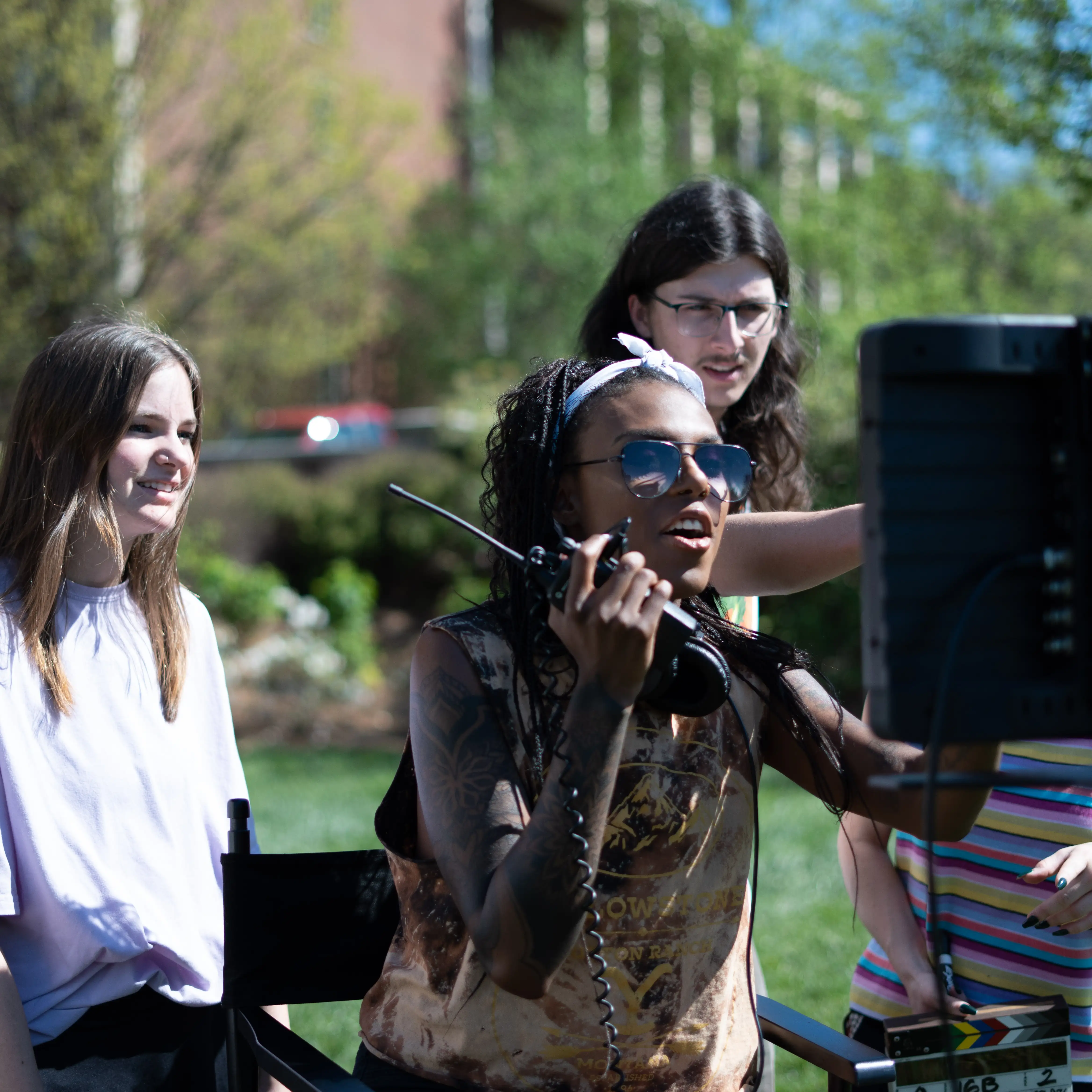 Students work together on an outdoor film set, one speaking into a walkie-talkie while monitoring a screen, with others gathered around offering input and watching closely.