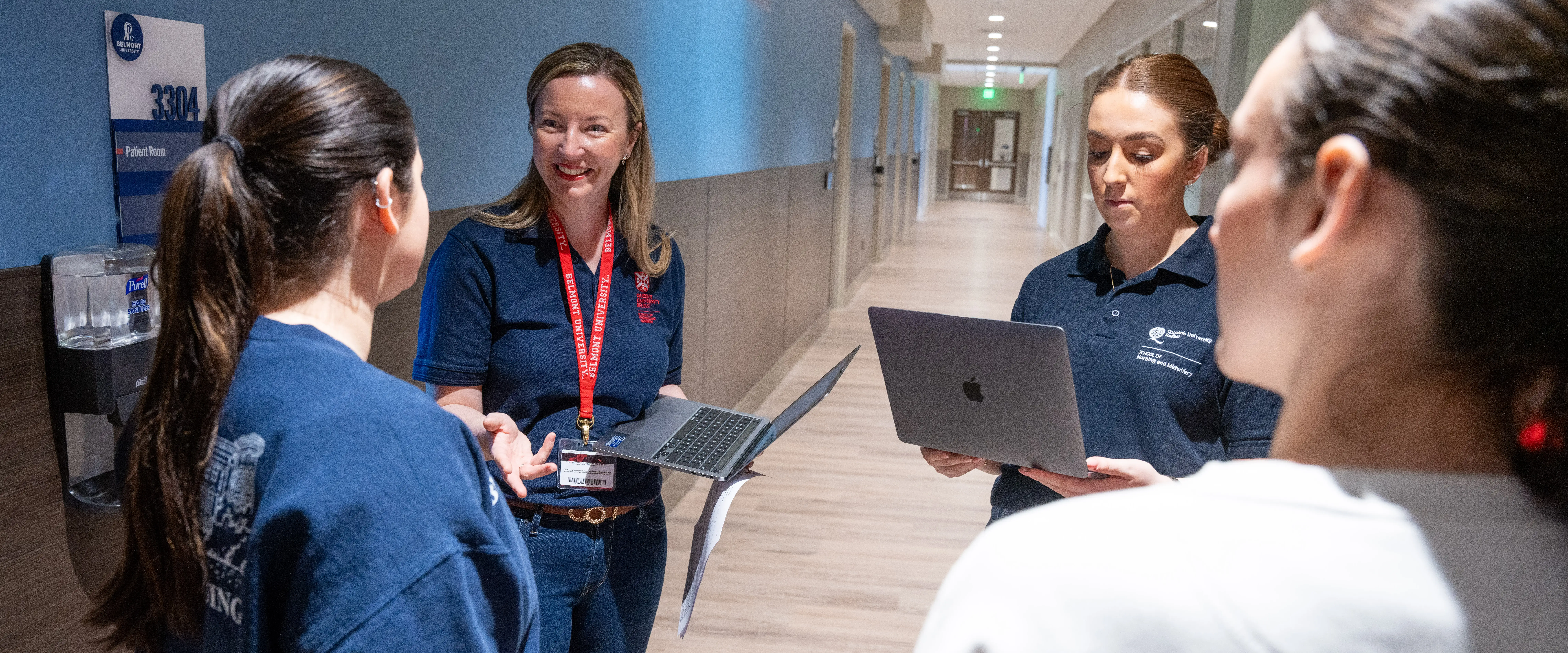Nursing faculty member in navy Belmont polo smiles while speaking with students in a hospital simulation hallway, holding a laptop.
