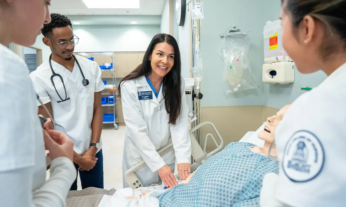 A group of healthcare students gather around a patient simulator in a clinical skills lab, with one student demonstrating a procedure. The setting is equipped with medical supplies and equipment, creating a realistic learning environment for hands-on training.