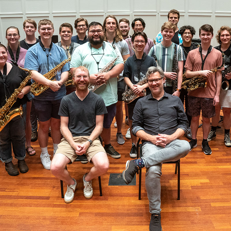 A group of students and professors stand smiling with their orchestra instruments.