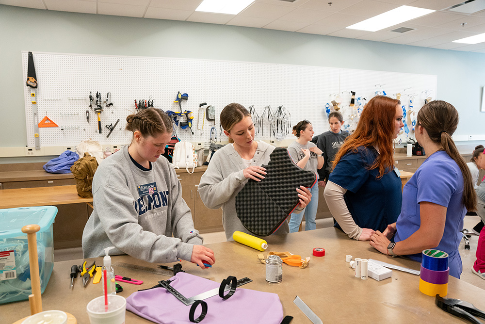 A group of OT students work on hands-on projects in a classroom equipped with tools and materials. One student in a gray sweatshirt is focused on assembling or modifying an item, while another holds a foam-padded structure. Others in scrubs and casual attire discuss their projects. The background features a pegboard with various tools and supplies, indicating a workshop or lab environment.