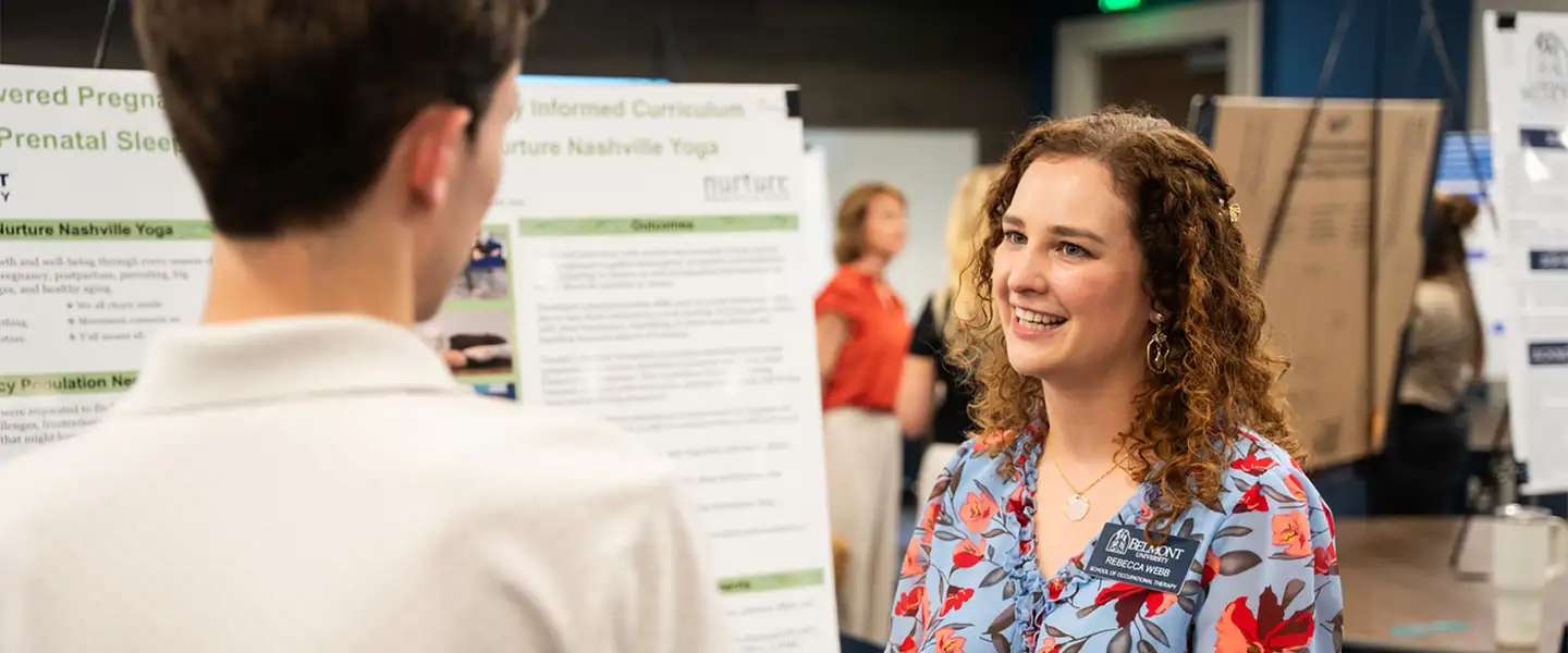 An occupational therapy student standing in front of her research poster at a conference, smiling and gesturing as she speaks with an attendee whose back is to the camera.