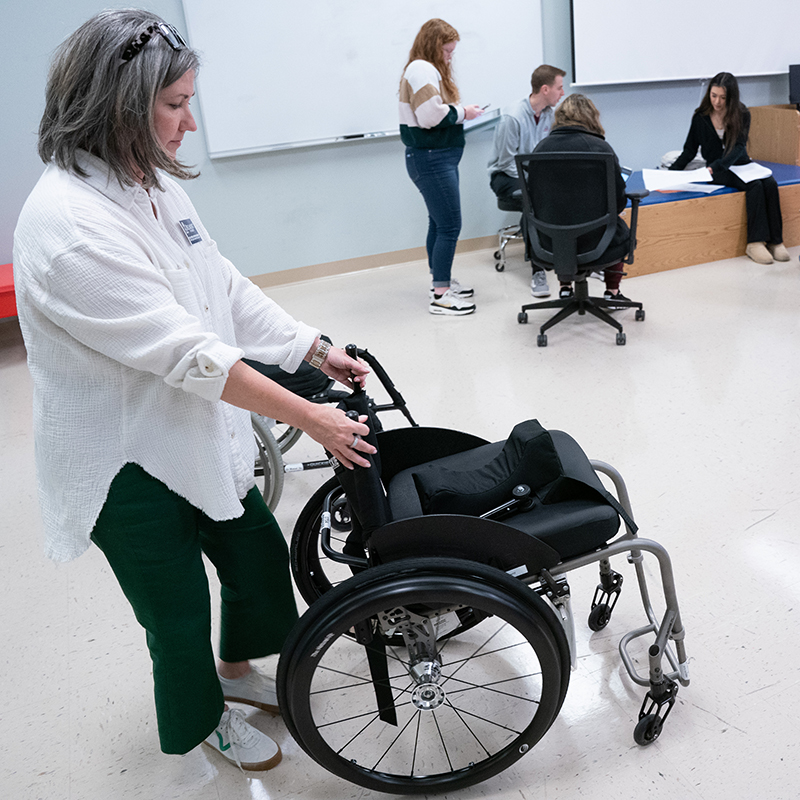 A faculty member guides students in a classroom, pushing a wheelchair while explaining its features. Other students are engaged in discussion in the background.