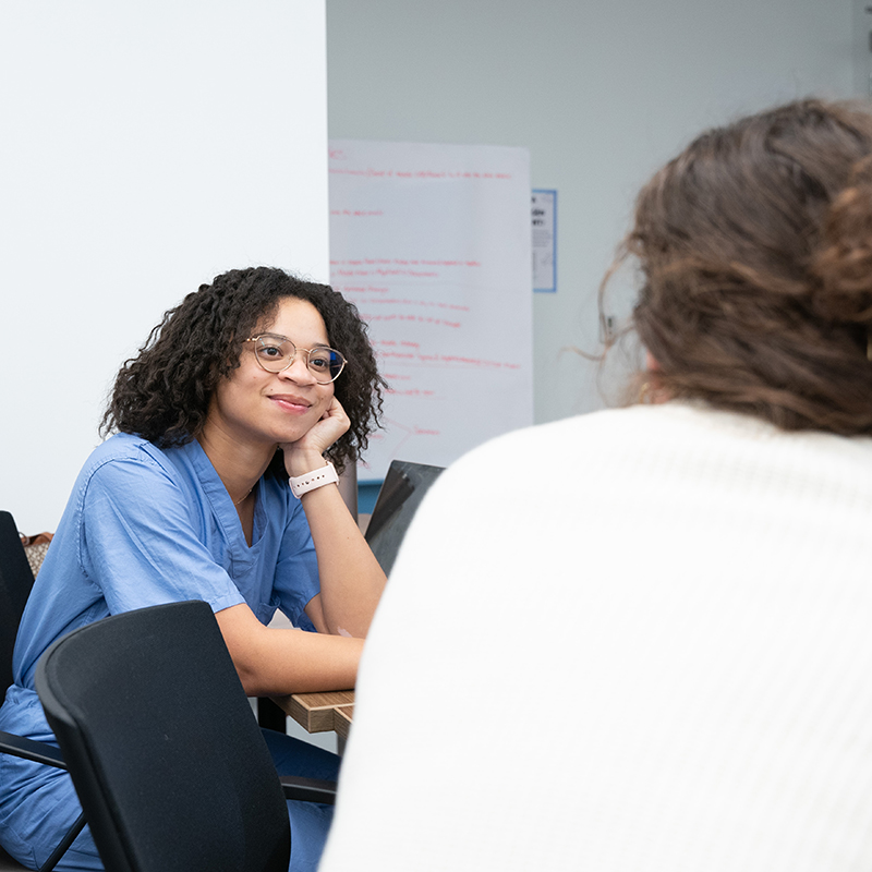 A student in blue scrubs smiles and listens attentively during a class discussion, while another student is visible in the background.  inline-mhc-classroom.jpg: A student in a navy polo gestures while giving a presentation in a classroom, with a digital screen visible in the background.