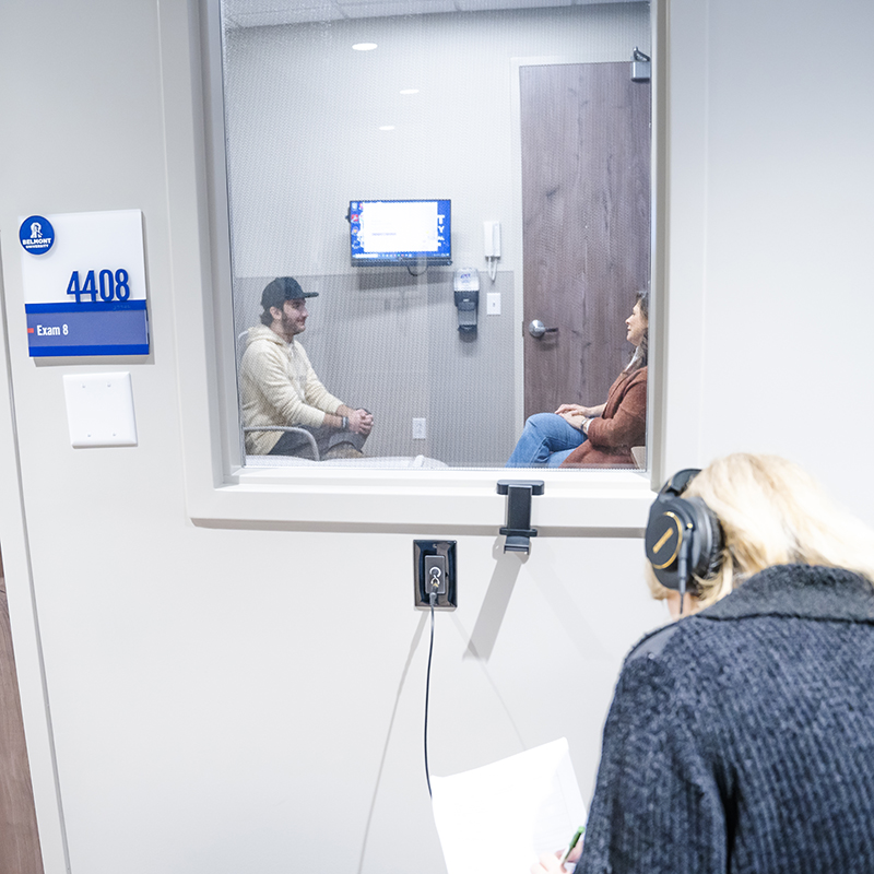 Two students are observed through a window as they participate in a counseling session simulation. A professor monitors from outside the room, wearing headphones.