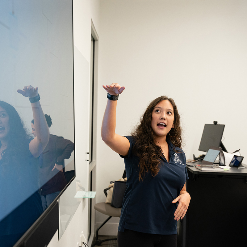 A student in a navy polo gestures while giving a presentation in a classroom, with a digital screen visible in the background.