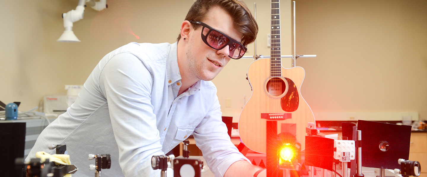 A male student wearing work goggles stands in front of a guitar and shines a red laser.