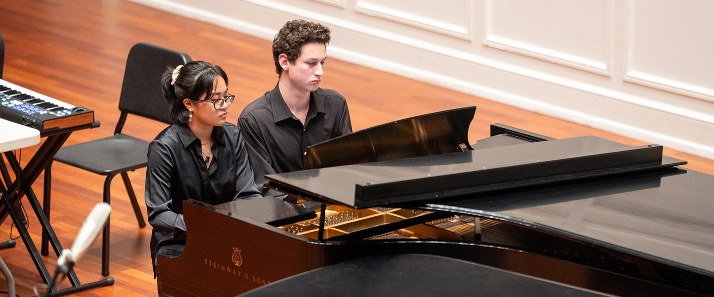 Two students, dressed in black, perform together on a grand piano during a recital. One plays the keyboard while the other plays the piano, both focused on their performance.