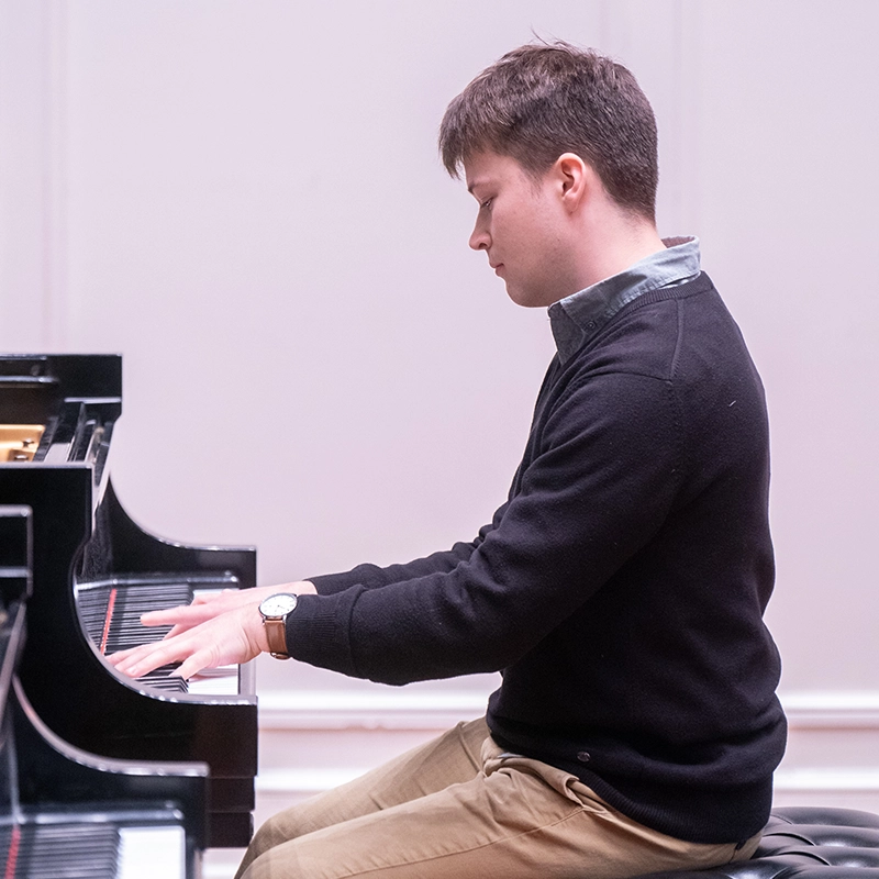 A student wearing a dark sweater and beige pants plays the piano, focused on his performance during a recital, with a Steinway piano visible in the frame.