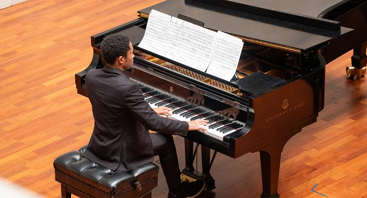 A pianist, dressed in a black suit, performs on a grand piano with sheet music in front of him. The reflection of his hands is visible on the piano's glossy surface.