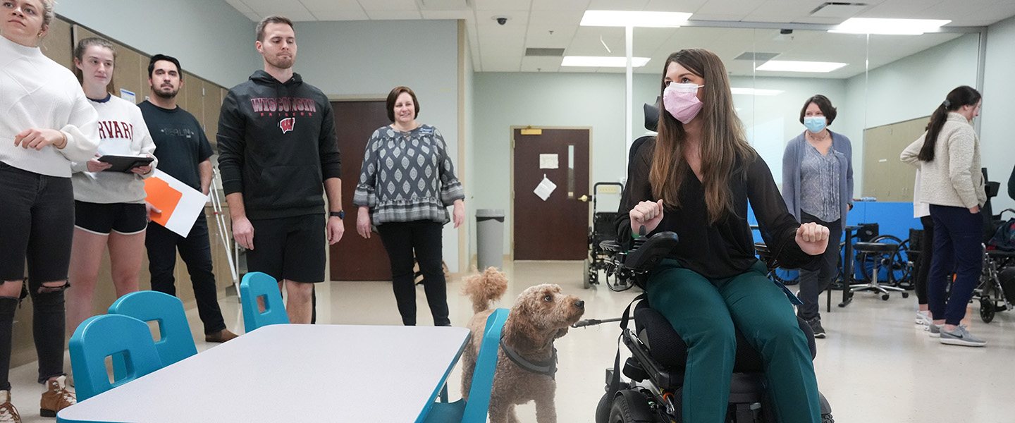 A masked female student is demonstrating using a wheel chair while her classmates and professor look on.